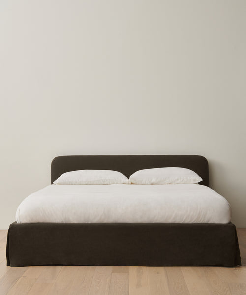 A minimalist Cove Bed with a dark upholstered headboard, dressed in crisp white Libeco linen and two pillows, sits on a light wooden floor against an off-white wall.
