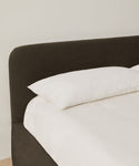 A neatly made Cove Bed with a dark upholstered headboard and crisp white Libeco linen rests against a plain, light-colored wall.