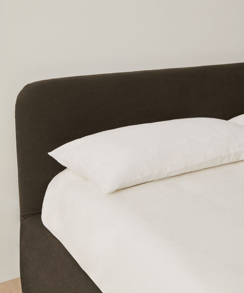 A neatly made Cove Bed with a dark upholstered headboard and crisp white Libeco linen rests against a plain, light-colored wall.