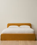 The Cove Bed showcases a mustard linen upholstered headboard and frame, accented with crisp white bedding and two pillows, set on a light wood floor beside a plain beige wall.