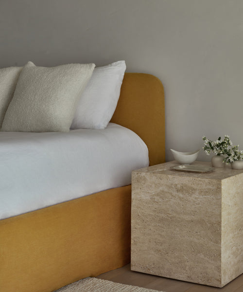 A minimal bedroom with a Cove Bed featuring a mustard linen headboard, white bedding, textured pillows, and a stone nightstand topped with small white dishes and fresh white flowers.