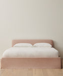 A minimalist bedroom showcases the Cove Bed with a pale pink linen headboard, crisp white Belgian Libeco Linen bedding, and two pillows, set against an off-white wall and light wood floor.
