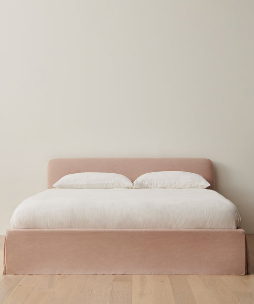 A minimalist bedroom showcases the Cove Bed with a pale pink linen headboard, crisp white Belgian Libeco Linen bedding, and two pillows, set against an off-white wall and light wood floor.
