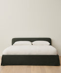 The Cove Bed, featuring a Belgian linen headboard, is dressed with two white pillows and a white duvet and placed on a light wooden floor against a plain, light-colored wall.