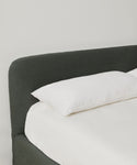 The Cove Bed features a dark upholstered headboard and a single white pillow on crisp white sheets, all set against a plain white wall.