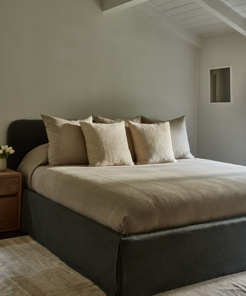 A minimalist bedroom with a Cove Bed dressed in beige linens and four pillows, a wooden nightstand holding a small vase of flowers, and neutral walls beneath a slanted ceiling.