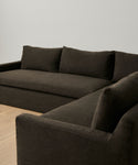 The Harbor Sectional, featuring plush cushions and a modern dark brown design, rests on a light wooden floor against a plain white wall in a minimalist room.