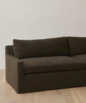A dark brown Harbor Sofa with linen upholstery and clean lines sits on a light wooden floor against a white wall. Only part of this handmade-in-Los Angeles piece, including one armrest and the backrest, is visible.