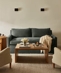 A cozy living room features the dark green linen-upholstered Harbor Sofa, a light throw blanket, a wooden coffee table with books, a candle, and white tulips, two beige armchairs, soft lighting, and cream-colored walls.