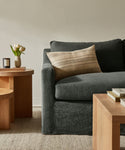A cozy living room corner highlights the Harbor Sofa with dark gray linen upholstery, paired with a striped beige pillow, light wood coffee tables, white tulips in a vase, a candle, and books on a neutral woven rug against a light wall.
