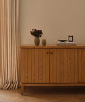 A wooden cabinet with vertical grooves stands against a cream wall. On top are dark flowers in a vase, a small ceramic vessel, books, and the Vail Glass Candle with a holiday scent. Cream curtains hang to the left.