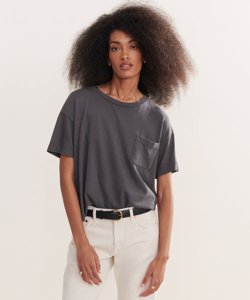 A woman with voluminous curly hair wears a Vintage Pocket Tee in charcoal, tucked into high-waisted light cream jeans with a black belt, standing against a plain light background.