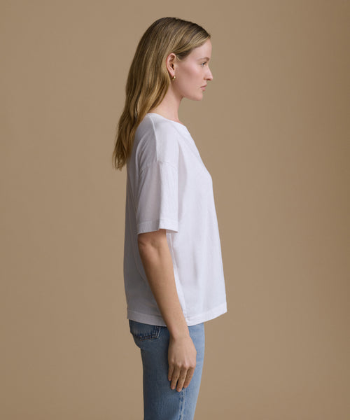 A woman in profile wearing a white short-sleeve crewneck t-shirt and light blue jeans, standing against a solid beige studio background.