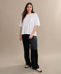 A woman with long brown hair and a neutral expression stands against a beige background, wearing a casual basic white Vintage Pocket Tee, black wide-leg pants, and brown flip-flops.