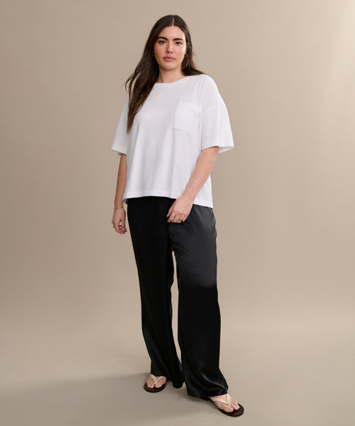 A woman with long brown hair and a neutral expression stands against a beige background, wearing a casual basic white Vintage Pocket Tee, black wide-leg pants, and brown flip-flops.