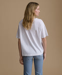 A woman is shown from the back wearing an oversized white short-sleeve t-shirt and light blue jeans, standing against a solid beige studio background.