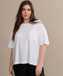 A woman with long brown hair stands in front of a beige background, wearing the Vintage Pocket Tee and black pants, looking at the camera with a neutral expression.