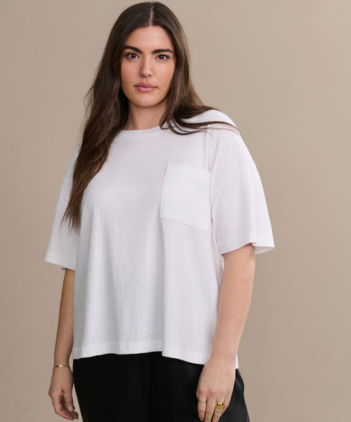 A woman with long brown hair stands in front of a beige background, wearing the Vintage Pocket Tee and black pants, looking at the camera with a neutral expression.