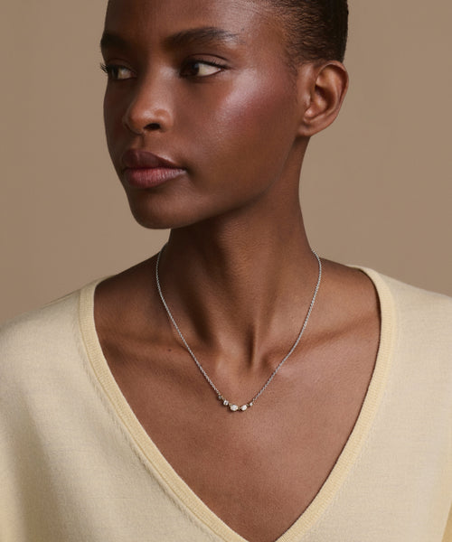 A woman with short hair wears a light yellow V-neck top and the Violette Diamond Necklace—three delicate white gold beads—looking to her left against a plain beige background for a subtle touch of fine jewelry.
