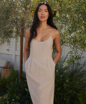 A woman stands outdoors by greenery and a white fence, wearing the sleeveless Rove Dress. This spring-summer style features thin straps, pockets, and Italian fabric as she looks confidently at the camera.