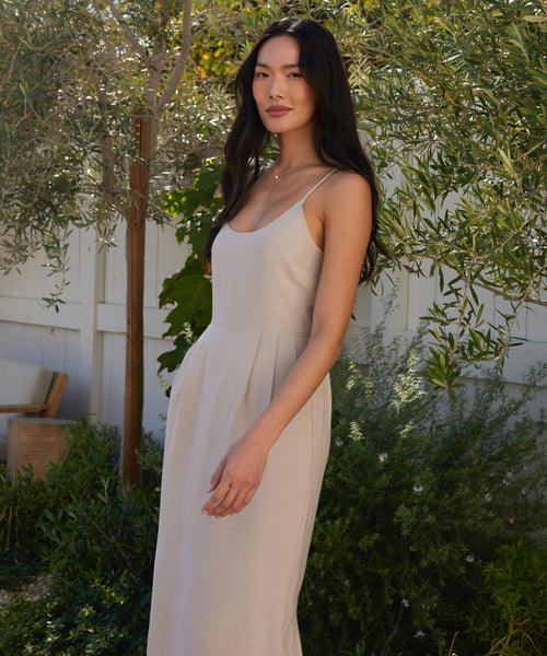 A woman with long dark hair stands outside by a white fence and green foliage, wearing the sleeveless light beige Rove Dress, made from fine Italian fabric, gazing calmly at the camera.