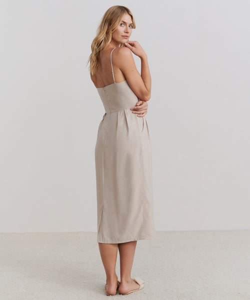 A woman stands on a light floor, facing slightly away and looking over her shoulder. She wears the sleeveless Rove Dress, a stylish choice for spring and summer, set against a plain, minimal backdrop.