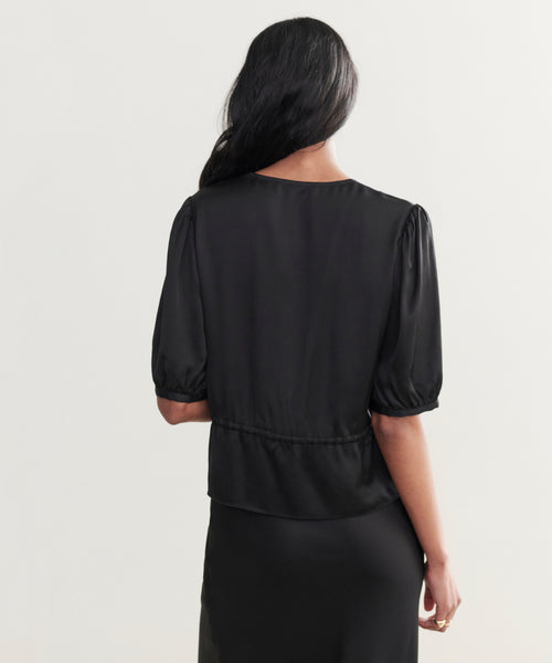 A woman stands facing away, wearing the Sawyer Blouse in black satin with puffed sleeves and a matching skirt. Her long dark hair drapes over her shoulder against a plain light background.
