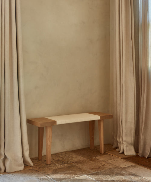 The Oak Day Bench, featuring a beige cushion, rests against a textured light beige wall with floor-length beige curtains nearby on stone tiles. Soft sunlight streams through the curtains.