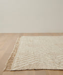 The Cabin Rug, cream-colored with Moroccan-inspired design and fringe on one edge, is placed on a light wooden floor against an off-white wall.