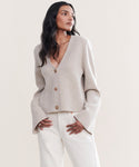 A woman with long dark hair wears the Chelsea Cardigan in light beige, featuring wide sleeves, paired with white pants. She stands against a plain light background, gazing to the side with one hand in her pocket.