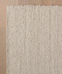 The Canyon Rug, a handmade, off-white wool piece from India with a chunky looped texture, is shown from above on a light wooden floor. The image captures a corner of this artisanal rug.