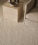 The Canyon Rug, a beige wool piece handmade in India, is placed beneath a stone square coffee table with decor, beside a light-upholstered armchair.
