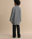 A person with short curly hair stands against a beige background, wearing a loose-fitting gray coat and Wool Jones Trouser, facing away from the camera.