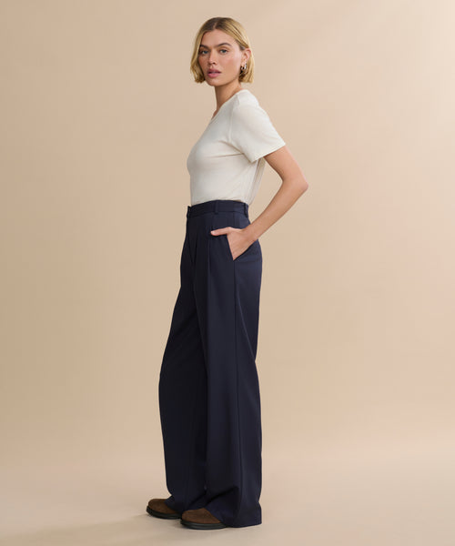 A woman stands in profile against a beige background, wearing a fitted short-sleeve white top, brown shoes, and navy Wool Jones Trousers. Her hands are in her pockets and she has short blonde hair.