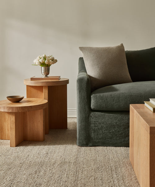 A modern living room with a dark green sofa, light gray pillow, wooden coffee and side tables, a small bowl, and a silver vase with white flowers on a textured Rug Swatch in beige.