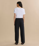 A woman with long wavy brown hair stands against a beige background, facing away. She wears a white short-sleeve top, black shoes, and the black Wool Paloma Pant featuring an elastic drawstring waist.