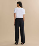 A woman with dark, side-parted hair stands with her back to the camera against a beige background, wearing a white short-sleeve t-shirt and black Wool Paloma Pant featuring an elastic drawstring waist.