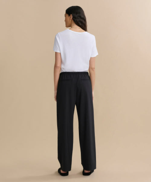 A woman with long wavy brown hair stands against a beige background, facing away. She wears a white short-sleeve top, black shoes, and the black Wool Paloma Pant featuring an elastic drawstring waist.