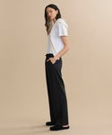 A woman stands in profile against a beige background, wearing a white t-shirt, the Wool Paloma Pant with an elastic drawstring waist, black slippers, and a white sweater draped over her shoulders. Her hands are in her pockets and her hair is loose.