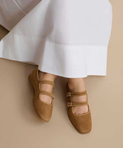 A person wearing a long white skirt and Wren Flats—light brown, double-strap Mary Janes with a rounded square toe—sits against a neutral beige background.