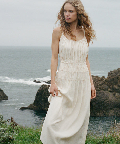 Woman in a cream wynn dress standing by the ocean with rocky cliffs.