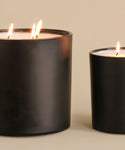 Two black candles are lit—an Extra Large Topanga Glass Candle with three flames on the left and a smaller one on the right, both hand-poured in Los Angeles and set against a neutral beige background.