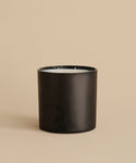 The Extra Large Topanga Glass Candle features a sleek black cylindrical design with three wicks. Hand-poured in Los Angeles, it is displayed on a beige surface with a matching beige background.