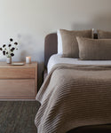 A modern bedroom showcases a neatly made Cove Bed in charcoal linen with textured brown pillows and a knitted beige blanket. A wooden nightstand holds a vase of dark flowers, candle, and tray, adding to the cozy, minimalist vibe.