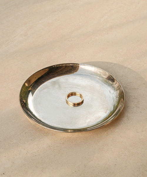 A single 14k solid gold James Band rests on a shiny, round silver plate atop a smooth beige surface, with soft natural light casting gentle shadows—a timeless piece from Jenni Kayne Jewelry.