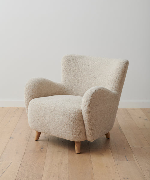 A chair in Bone Sherpa fabric with textured, cream upholstery and curved armrests stands on light wooden legs atop a pale floor, reflecting the cozy appeal of our Aspen Sofa collection. Featured fabric: Fabric Swatch Bone Sherpa.