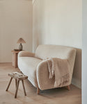 A Fabric Swatch Bone Sherpa sample complements a cozy Aspen Sofa with a fringed throw in a minimalist living room featuring light wood floors, a small wooden stool, and lamp against a neutral wall.