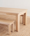 A light wood dining table and the Dining Bench, both on a woven beige rug, set against a plain white wall.