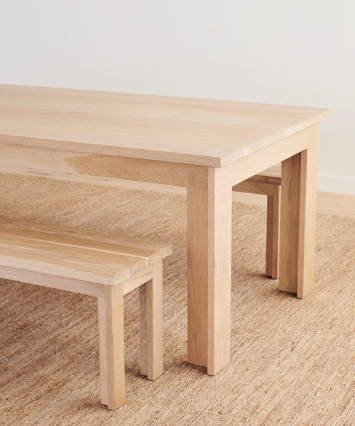 A light wood dining table and the Dining Bench, both on a woven beige rug, set against a plain white wall.