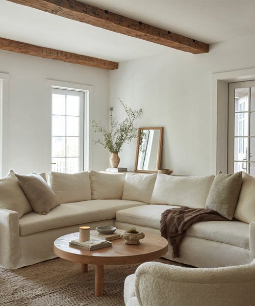A cozy living room features the Harbor Sectional in Belgian flax linen, neutral pillows, a brown throw, a round wood coffee table, and a vase of greenery. Wooden ceiling beams and large windows create a bright, inviting atmosphere.